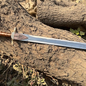 Op de afbeelding: Een decoratief zwaard met een zilveren kling en een bronzen gevest. Het gevest heeft een gedetailleerd ontwerp en de handgreep is donkerbruin. Het zwaard ligt op een verweerde boomstam, wat wijst op een rustiek of historisch thema.