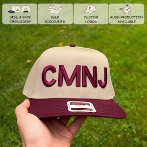 May include: A beige baseball cap with a maroon flat brim and the letters CMNJ embroidered in maroon on the front panel. A rectangular sticker with the text OTTO CLASSIC OSFM MID PROFILE MAXIMUM COMFORT is attached to the brim.