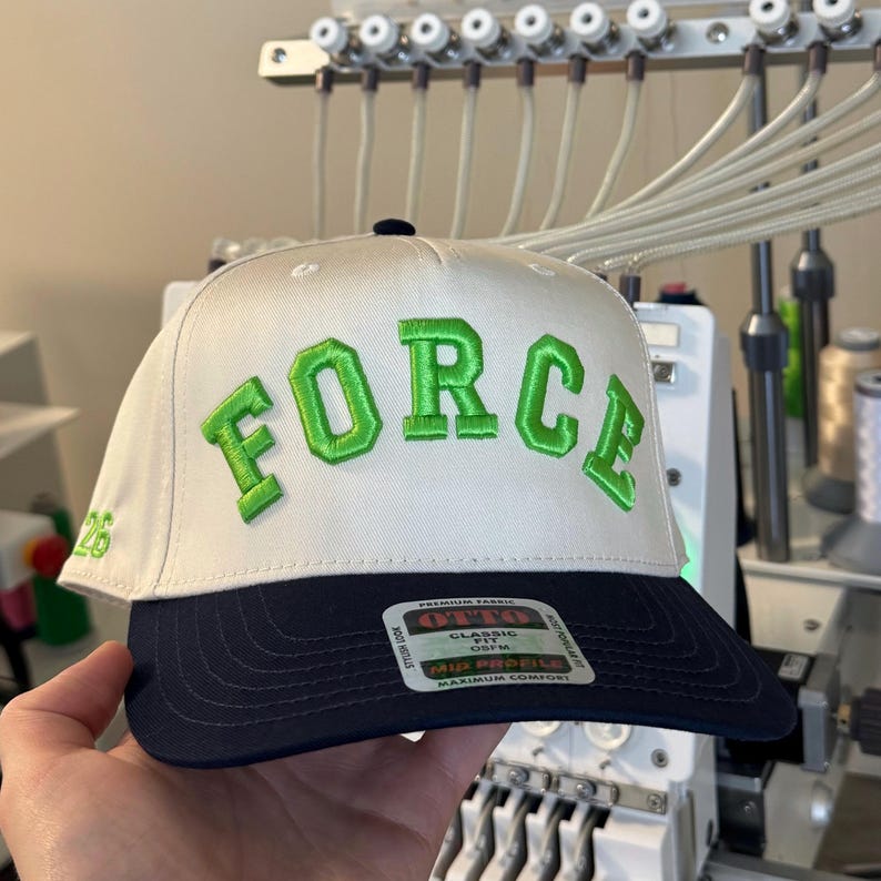 May include: A white and navy blue baseball cap with the word "FORCE" embroidered in green across the front. The number "26" is embroidered on the side. The cap has a flat brim and a structured fit.