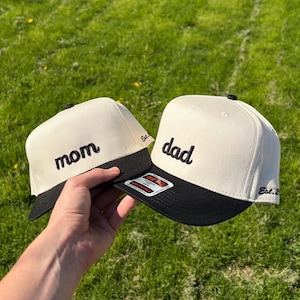 May include: Two baseball caps with a cream-colored crown and a black brim. One cap has the word "mom" embroidered in black, and the other says "dad". Both caps have a small black button on top and the text "Est. 2" on the side.