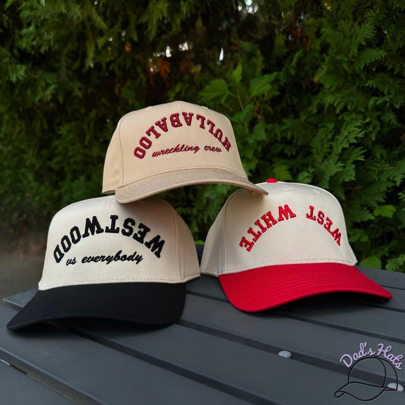 May include: Three baseball caps with different designs. One is tan with red text that says "HULLABALOO wrecking crew". Another is off-white with black text that says "WESTWOOD us everybody". The third is off-white with a red brim and red text that says "WHITE WEST".