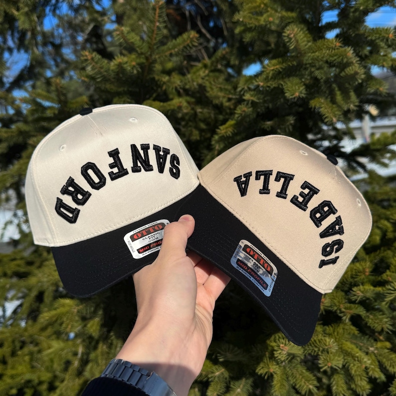 May include: Two baseball caps with black brims and the names "SANFORD" and "ISABELLA" embroidered in black on the front. The caps have a cream-colored crown. The caps are held up against a backdrop of green pine needles.
