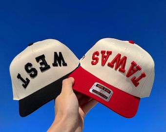 May include: Two baseball caps with the text "WEST" and "TAWAS". The "WEST" cap has a black brim and black lettering, while the "TAWAS" cap has a red brim and red lettering. Both caps have a cream-colored crown.