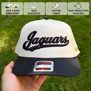 May include: A two-tone baseball cap featuring an off-white crown with the word Jaguars embroidered in black script across the front. The cap has a solid black brim and includes a rectangular sticker on the brim with the text Otto Premium Fabric.
