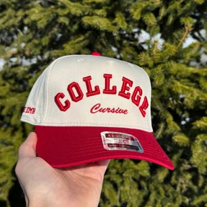 May include: A white and red baseball cap with the word "COLLEGE" embroidered in red block letters and "Cursive" in red script. The cap has a red brim and a small red button on top.
