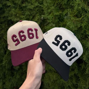 May include: Two baseball caps with the year "1995" embroidered on the front. One cap has a tan crown and a burgundy brim, with burgundy text. The other cap has a cream crown and a black brim, with black text.