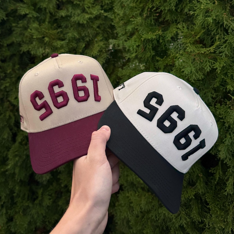 May include: Two baseball caps with the year "1995" embroidered on the front. One cap has a tan crown and a burgundy brim, with burgundy text. The other cap has a cream crown and a black brim, with black text.