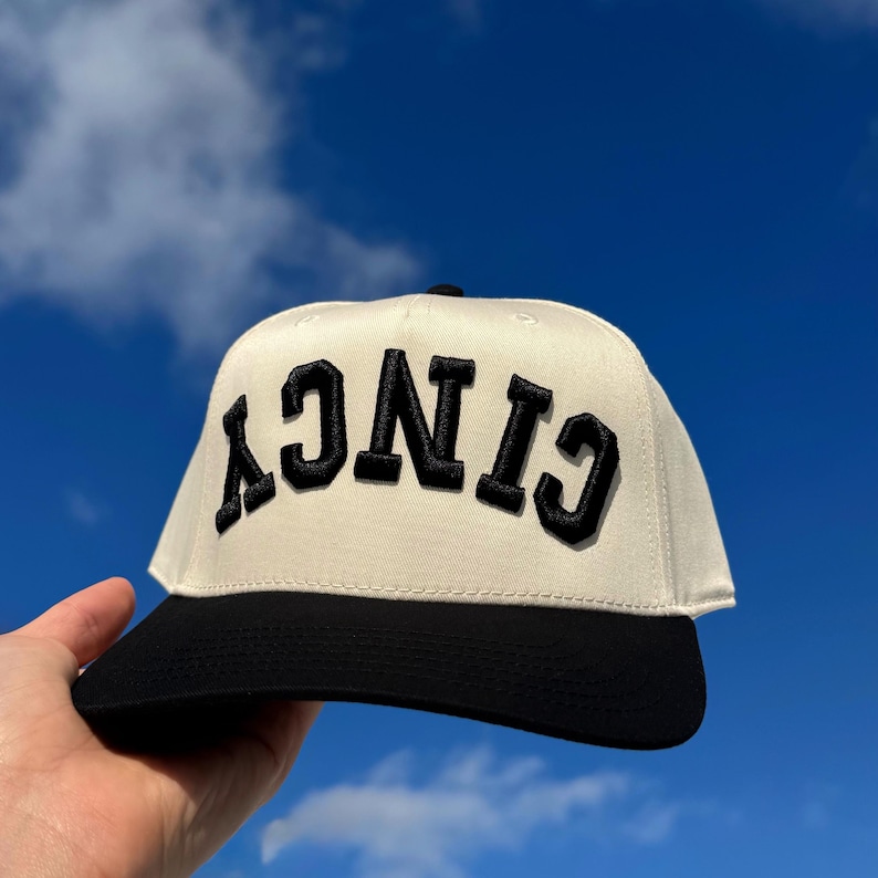 May include: Cream-colored baseball cap with the word "Cincy" embroidered in bold, black letters. The cap features a black brim and a classic, structured design. The background is a bright blue sky with fluffy white clouds.