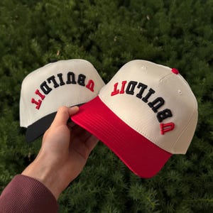 May include: Two baseball caps with the word "BUILD IT" embroidered on the front. One cap has a red brim and the other has a black brim. The caps are cream-colored with red and black lettering.