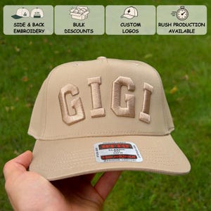 May include: A tan baseball cap featuring the word GIGI embroidered in large, raised, matching tan letters across the front panels. The cap includes a sticker on the brim with the text OTTO CLASSIC FIT OSFM and a mid-profile design.