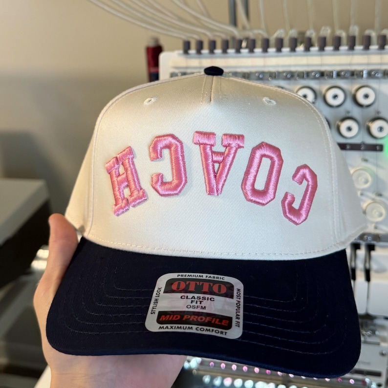 May include: A white and navy blue baseball cap with the word "COACH" embroidered in pink. The cap has a curved brim and a black button on top. The tag reads "OTTO CLASSIC FIT OSFM MID PROFILE MAXIMUM COMFORT".