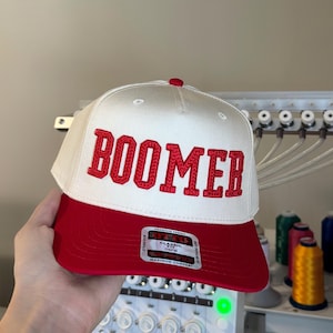 May include: A white and red baseball cap with the word "BOOMER" embroidered in red block letters. The cap has a red brim and a small red button on top. The cap has a "Classic Fit" label.