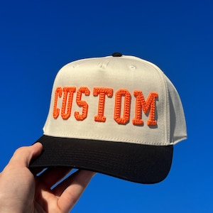 May include: A cream-colored baseball cap with the word "CUSTOM" embroidered in orange block letters. The cap has a black brim and a black button on top. The background is a bright blue sky.