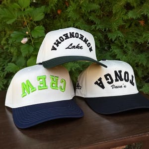May include: Three baseball caps with white crowns and colored brims are displayed. One cap has green embroidery that says "CREW", another has black embroidery that says "TONGA Vava'u", and the third has black embroidery that says "RONKONKOMA Lake".