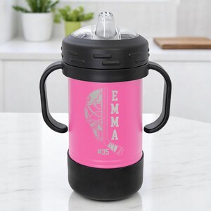 May include: A pink insulated tumbler with a black lid, handle, and base. The tumbler features a hockey-themed design with the name "EMMA" and the number "35" etched on the side. The tumbler has a clear, flip-top spout.
