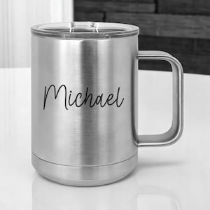 May include: A stainless steel travel mug with a clear lid and a large handle. The name "Michael" is written in black script on the side of the mug. The mug is designed to keep beverages hot or cold.