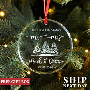 Op de afbeelding: Een heldere, ronde kerstversiering met de tekst "OUR FIRST CHRISTMAS Mr & Mrs" en de namen "Mark & Donna" met de datum "07.21.2024". De versiering heeft een rood lint om op te hangen en heeft een ontwerp van drie dennenbomen.