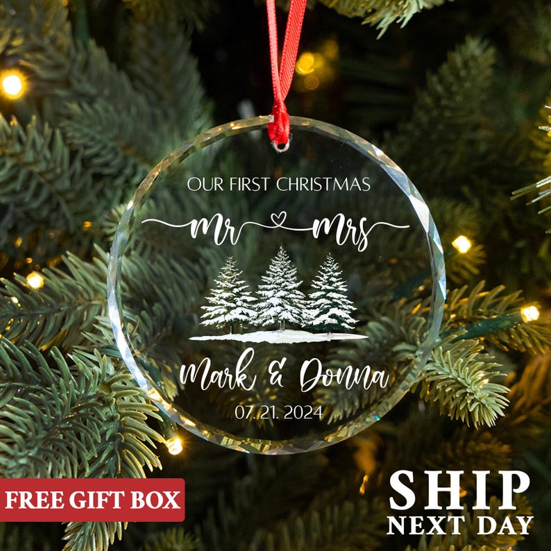 Op de afbeelding: Een heldere, ronde kerstversiering met de tekst "OUR FIRST CHRISTMAS Mr & Mrs" en de namen "Mark & Donna" met de datum "07.21.2024". De versiering heeft een rood lint om op te hangen en heeft een ontwerp van drie dennenbomen.