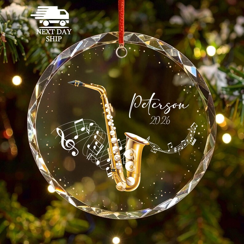 May include: A circular, faceted glass ornament featuring a gold saxophone illustration, musical notes, and the text "Peterson 2026" printed in white script. The ornament hangs from a red ribbon and has a metallic loop at the top for suspension.