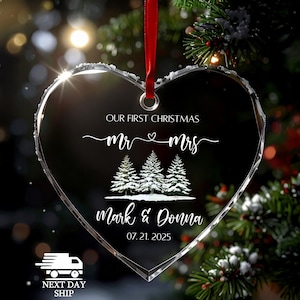 Op de afbeelding: Een heldere, hartvormige kerstversiering met een rood lint. De versiering bevat de tekst "OUR FIRST CHRISTMAS", "Mr & Mrs", drie met sneeuw bedekte dennenbomen en de namen "Mark & Donna" met de datum "07.21.2025".