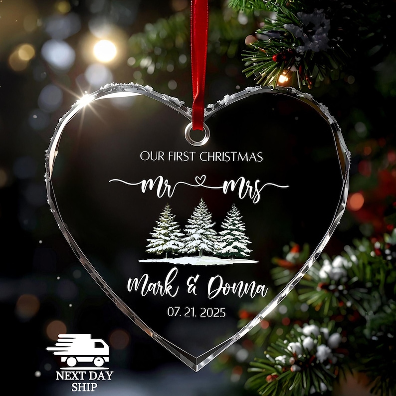 Op de afbeelding: Een heldere, hartvormige kerstversiering met een rood lint. De versiering bevat de tekst "OUR FIRST CHRISTMAS", "Mr & Mrs", drie met sneeuw bedekte dennenbomen en de namen "Mark & Donna" met de datum "07.21.2025".