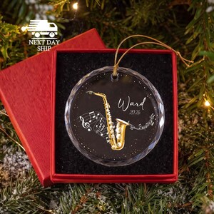 May include: A round, clear glass ornament with a faceted edge, featuring a gold saxophone illustration, musical notes, and the text "Ward 2026" in white script. The ornament hangs by a gold cord and is presented in a red gift box.