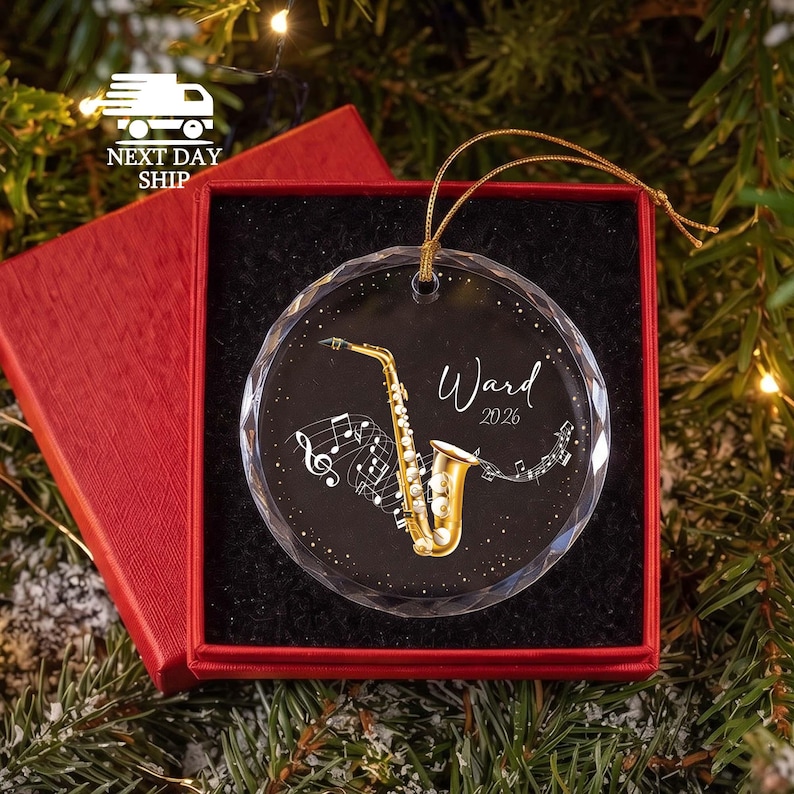 May include: A round, clear glass ornament with a faceted edge, featuring a gold saxophone illustration, musical notes, and the text "Ward 2026" in white script. The ornament hangs by a gold cord and is presented in a red gift box.