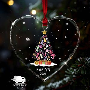 May include: A heart-shaped clear acrylic ornament hanging from a red ribbon. The center features a Christmas tree graphic composed of various hair styling tools and beauty products, topped with a gold star and sitting on colorful gift boxes. The name EVELYN and the year 2026 are printed in white below the tree.