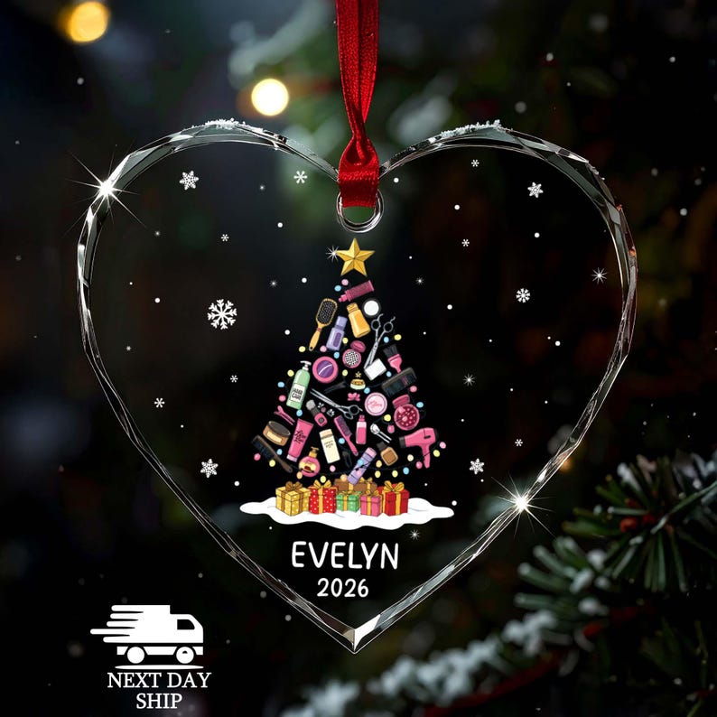 May include: A heart-shaped clear acrylic ornament hanging from a red ribbon. The center features a Christmas tree graphic composed of various hair styling tools and beauty products, topped with a gold star and sitting on colorful gift boxes. The name EVELYN and the year 2026 are printed in white below the tree.