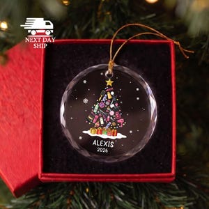 May include: A round, clear glass ornament featuring a black background with a Christmas tree graphic composed of various hair styling tools and beauty products. The text ALEXIS 2026 is printed in white at the bottom, and it hangs by a gold cord.