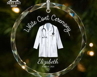May include: A clear, round ornament with a white lab coat and stethoscope design. The text "White Coat Ceremony" and the name "Elizabeth" are displayed, along with the date June 03, 2025, and a heart symbol. A silver string is attached for hanging.