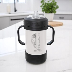 May include: A white and black sippy cup with a hockey-themed design. The cup features a hockey mask and stick graphic, the name "DAVID", and the number "07". It has a black lid, handles, and base.