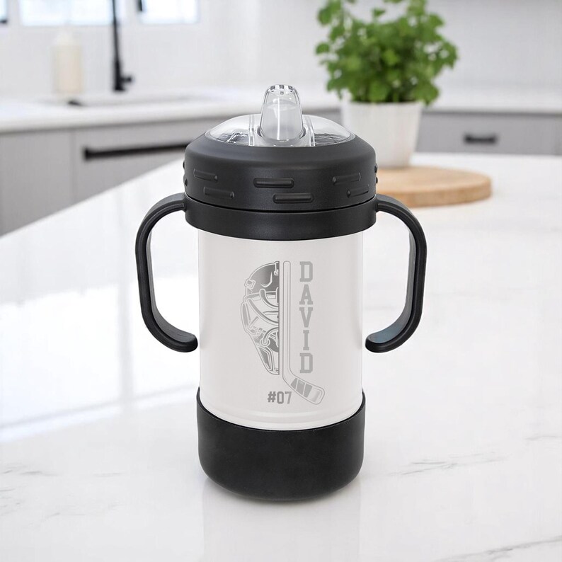 May include: A white and black sippy cup with a hockey-themed design. The cup features a hockey mask and stick graphic, the name "DAVID", and the number "07". It has a black lid, handles, and base.