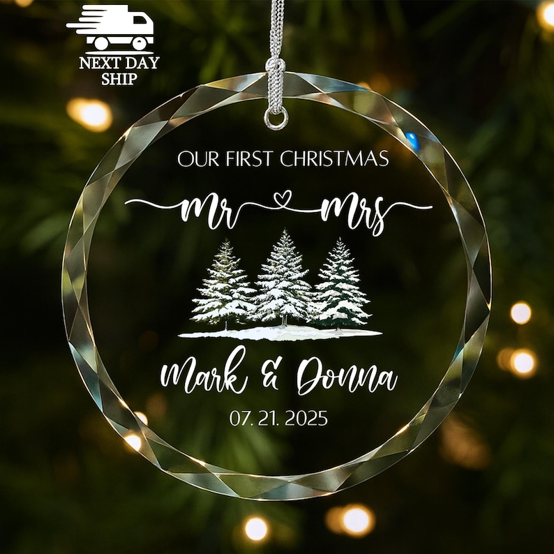Op de afbeelding: Transparante, ronde kerstversiering met een facetrand. De versiering heeft de tekst "OUR FIRST CHRISTMAS" boven "mr & mrs" en een afbeelding van drie met sneeuw bedekte dennenbomen. Daaronder staan de namen "Mark & Donna" en de datum "07.21.2025".