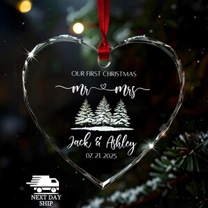 Op de afbeelding: Hartvormige kerstversiering van helder glas met een rood lint. De versiering heeft de tekst "OUR FIRST CHRISTMAS Mr & Mrs" en de namen "Jack & Ashley" met de datum "07.21.2025". Drie met sneeuw bedekte dennenbomen zijn ook afgebeeld.