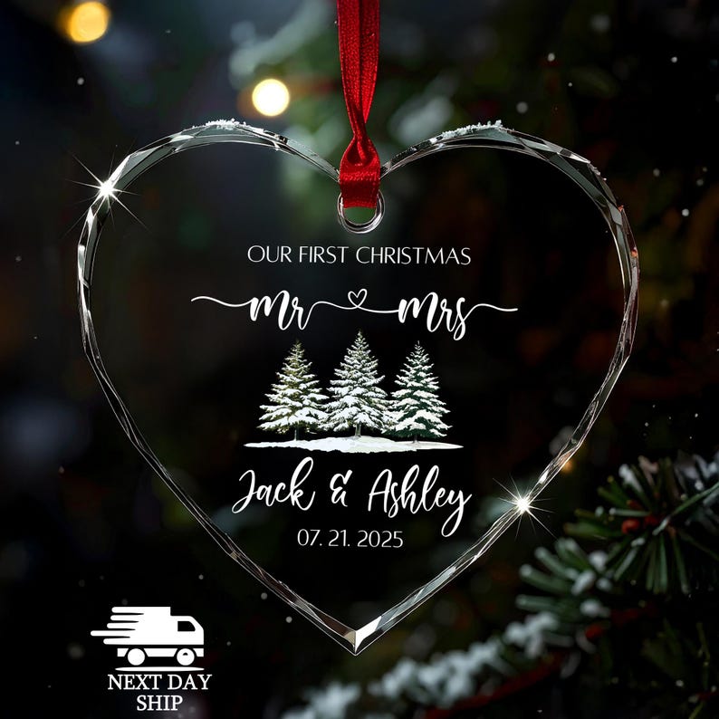 Op de afbeelding: Hartvormige kerstversiering van helder glas met een rood lint. De versiering heeft de tekst "OUR FIRST CHRISTMAS Mr & Mrs" en de namen "Jack & Ashley" met de datum "07.21.2025". Drie met sneeuw bedekte dennenbomen zijn ook afgebeeld.