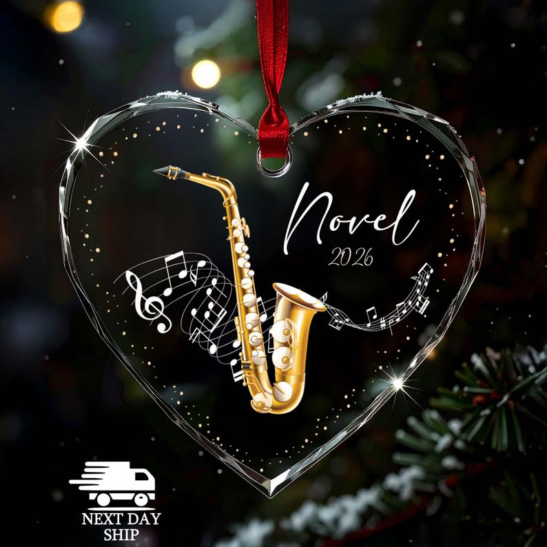 May include: A heart-shaped clear acrylic ornament featuring a gold saxophone illustration, white musical notes, and the text "Novel 2026" in a script font. The ornament is suspended by a red ribbon and has a faceted edge design.