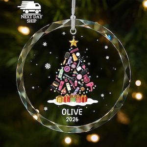 May include: A circular clear glass ornament featuring a Christmas tree graphic composed of various hair styling tools and beauty products. The tree is topped with a gold star and sits above colorful gift boxes. Text reads OLIVE 2026.