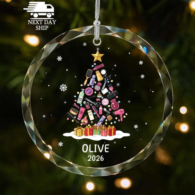 May include: A circular clear glass ornament featuring a Christmas tree graphic composed of various hair styling tools and beauty products. The tree is topped with a gold star and sits above colorful gift boxes. Text reads OLIVE 2026.