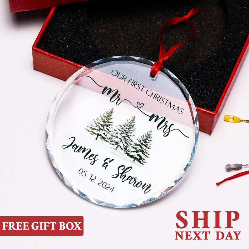 Op de afbeelding: Een heldere, ronde kerstversiering met een rood lint. De versiering heeft de tekst "OUR FIRST CHRISTMAS Mr & Mrs" boven een ontwerp van drie dennenbomen. Daaronder staat de tekst "James & Sharon 05.12.2024". De versiering zit in een rode geschenkverpakking.