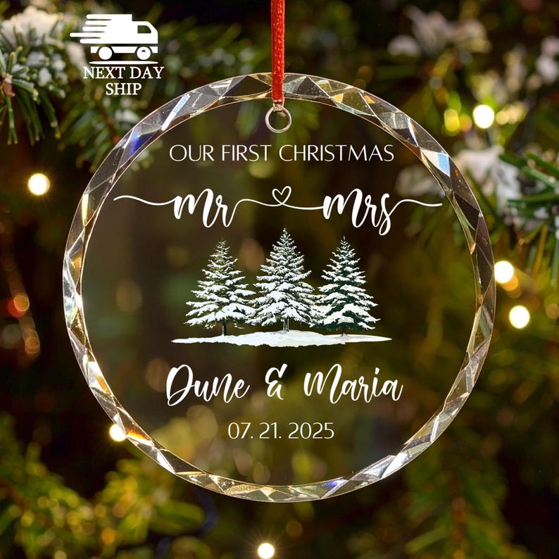 Op de afbeelding: Een heldere, ronde kerstbal met een rood lint. De ornament toont de tekst "OUR FIRST CHRISTMAS" boven "Mr & Mrs" met een hartsymbool. Daaronder staan drie met sneeuw bedekte dennenbomen en de namen "Dune & Maria" met de datum "07.21.2025".