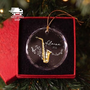 May include: A circular clear glass ornament featuring a gold saxophone illustration, white musical notes, and the text Alaina 2026. The ornament has a gold hanging cord and is presented in a black-lined red gift box for holiday display.