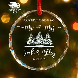 Op de afbeelding: Een heldere, facetgeslepen glazen kerstbal met een rood lint. De ornament bevat de tekst "OUR FIRST CHRISTMAS Mr & Mrs" boven drie met sneeuw bedekte dennenbomen en de namen "Jack & Ashley" met de datum "07.21.2025".