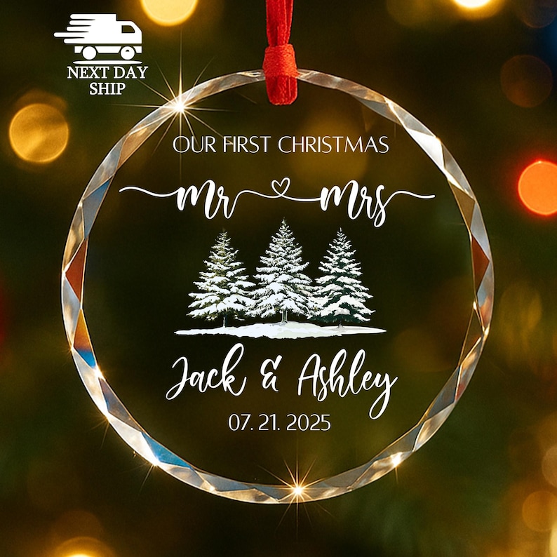 Op de afbeelding: Een heldere, facetgeslepen glazen kerstbal met een rood lint. De ornament bevat de tekst "OUR FIRST CHRISTMAS Mr & Mrs" boven drie met sneeuw bedekte dennenbomen en de namen "Jack & Ashley" met de datum "07.21.2025".