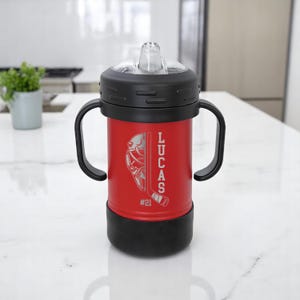 May include: Red insulated sippy cup with black handles and lid. The cup features a hockey-themed design with the name "LUCAS" and the number "#21" etched on the side. The cup has a clear, flip-top spout.