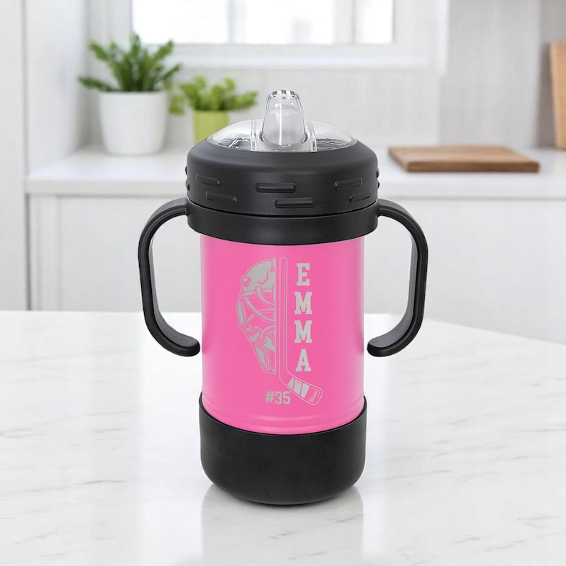 May include: A pink insulated tumbler with a black lid, handle, and base. The tumbler features a hockey-themed design with the name "EMMA" and the number "35" etched on the side. The tumbler has a clear, flip-top spout.
