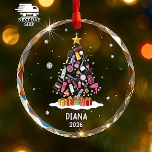 May include: A circular clear glass ornament featuring a printed Christmas tree design composed of various hair styling tools and beauty products. The tree is topped with a gold star and sits above colorful gift boxes with the name DIANA and 2026.