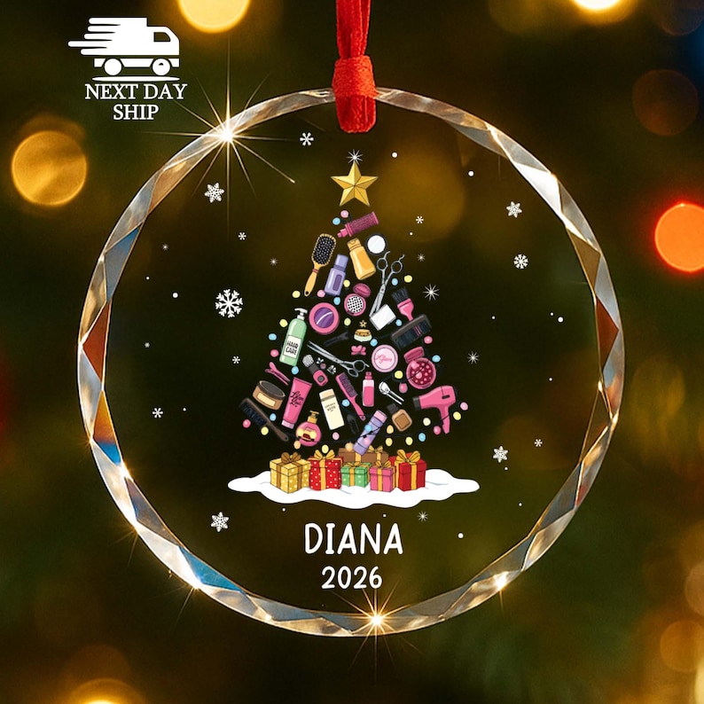 May include: A circular clear glass ornament featuring a printed Christmas tree design composed of various hair styling tools and beauty products. The tree is topped with a gold star and sits above colorful gift boxes with the name DIANA and 2026.