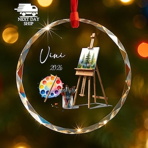 May include: A circular glass ornament with a faceted edge and a red ribbon hanger. The design features a wooden easel holding a forest painting, a colorful paint palette, and a jar of brushes. The text Vini and 2026 are printed on the surface.