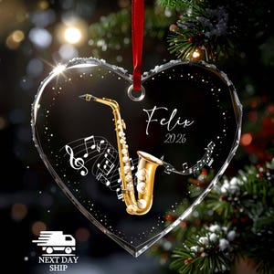 May include: A heart-shaped clear acrylic ornament featuring a gold saxophone illustration, white musical notes, and the text "Felix" above "2026". The ornament is suspended by a red ribbon and has a decorative border with small white dots.